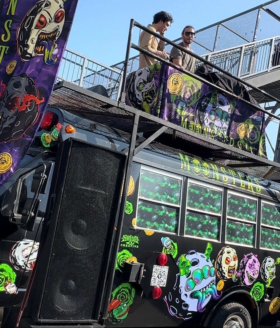 Decorated Moonsters stage bus with colorful designs and text, featuring a speaker on the side and 2 DJs performing on the stage.
