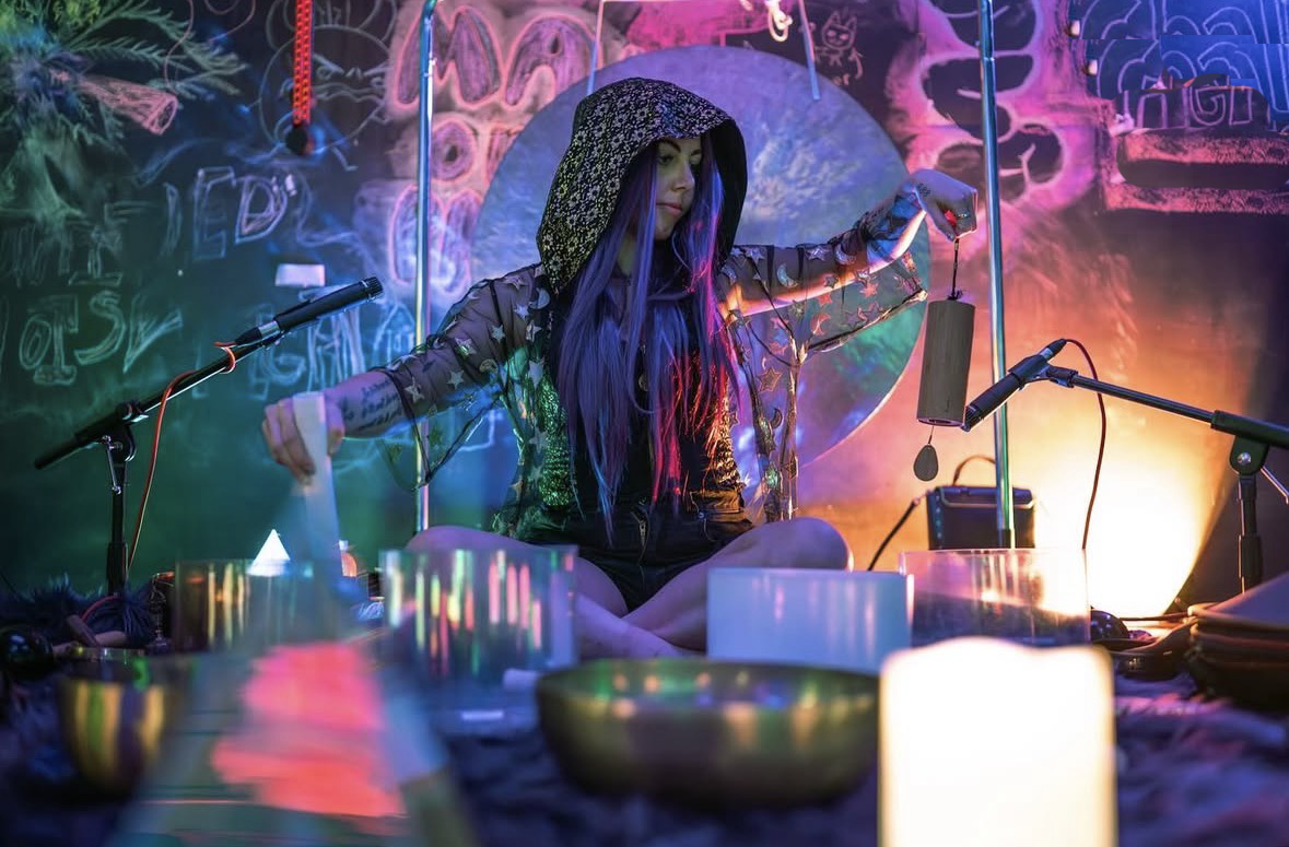 Female KT The Alchemist in a hooded outfit playing  instrument with colorful lights and decorations in the background