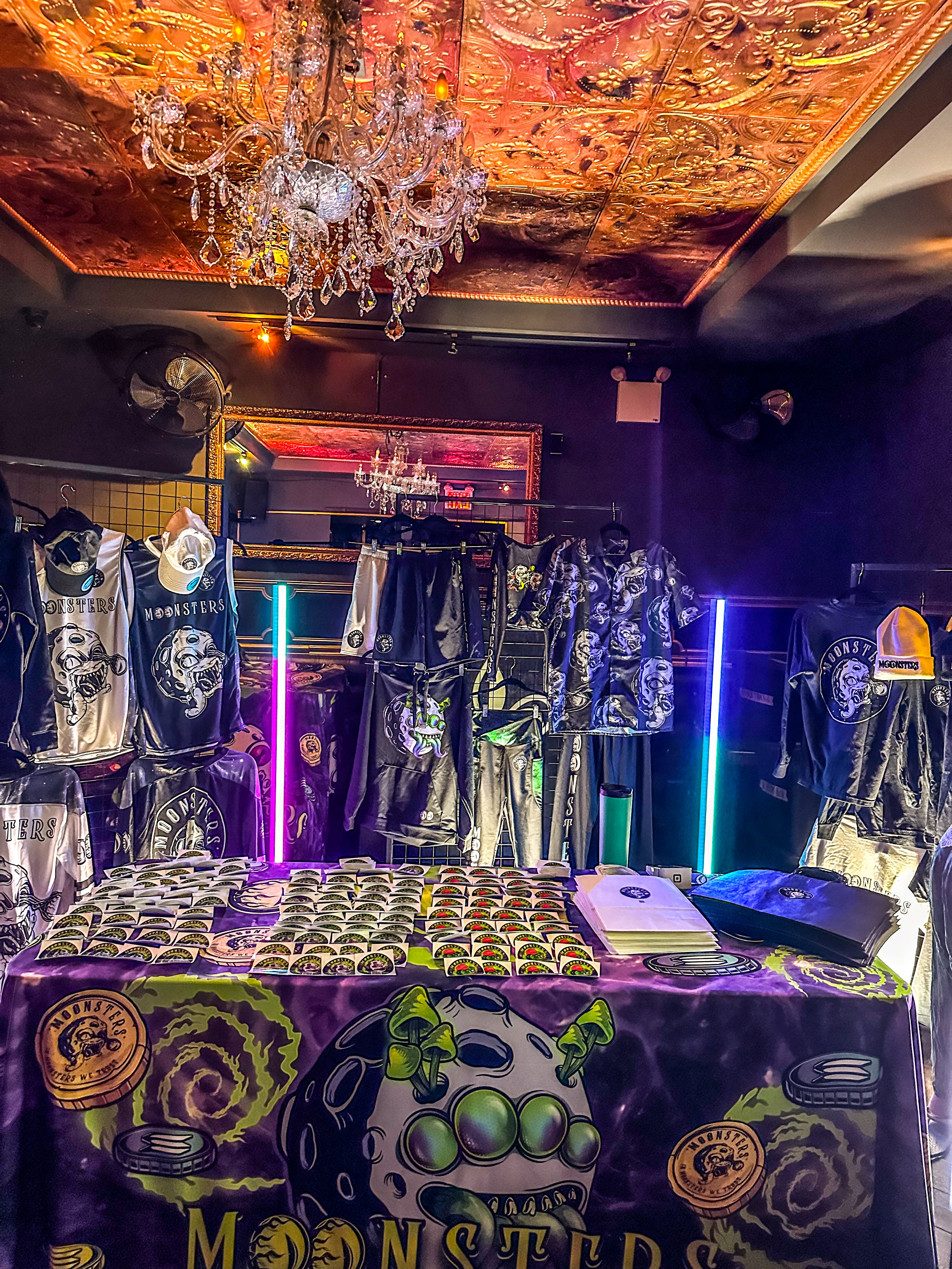 Display of Moonsters merchandise with a chandelier and colorful lighting in the background at hotel Chantelle in Manhattan for the Moonsters NFT NYC event.