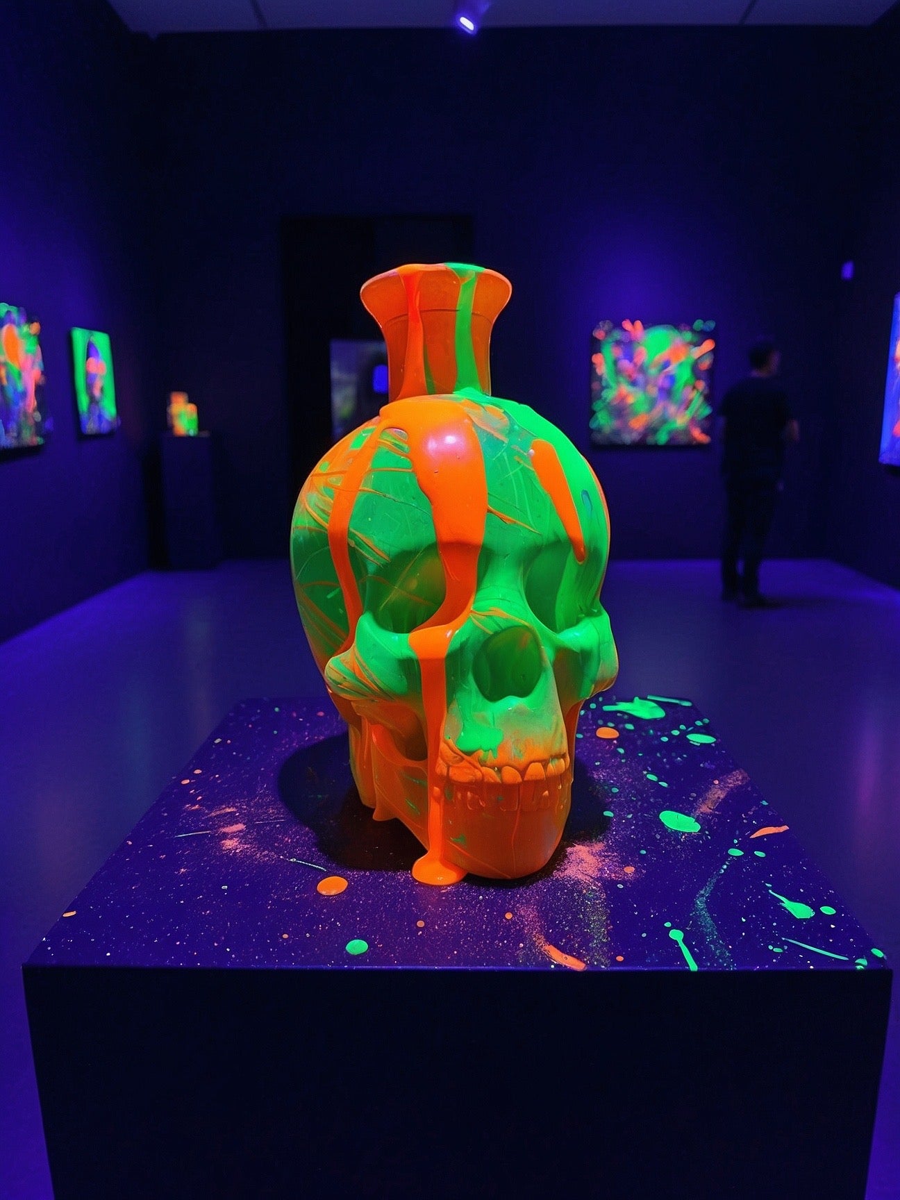 Neon orange and green skull sculpture with paint drips, displayed in dark gallery with glowing artwork.